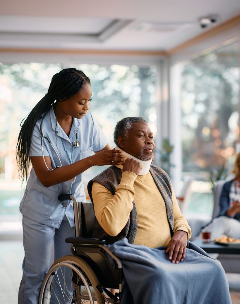 African American nurse putting cervical collar on senior man's neck at nursing home.