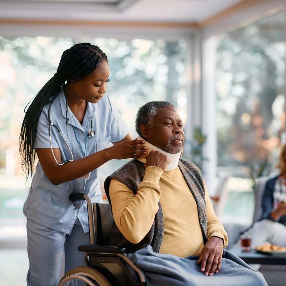 African American nurse putting cervical collar on senior man's neck at nursing home.